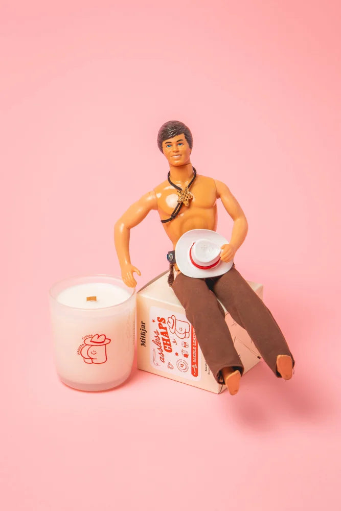 A vintage action figure with dark hair and brown pants sits on a box beside a saddle leather coconut soy candle