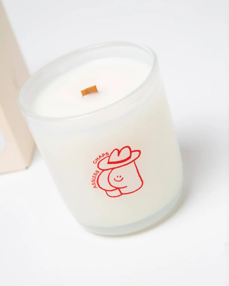 A frosted glass coconut soy candle jar with white wax, wooden wick, and a red cartoon cowboy hat for saddle leather fans