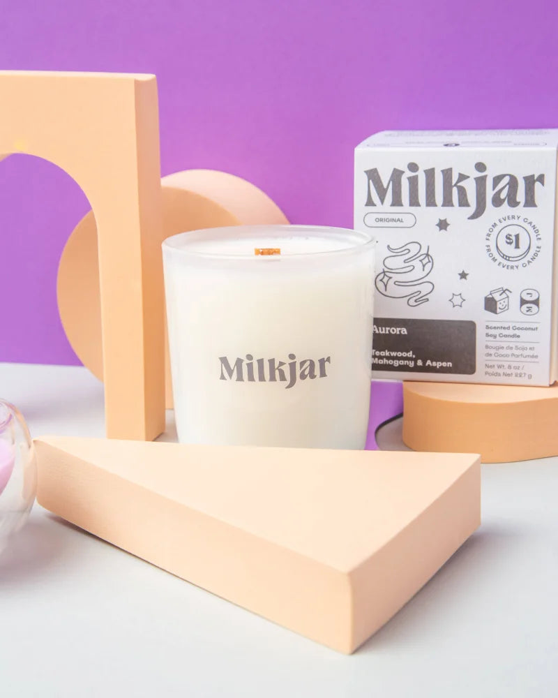A white aurora mahogany teakwood and aspen coconut soy candle in a clear glass jar with brand name visible