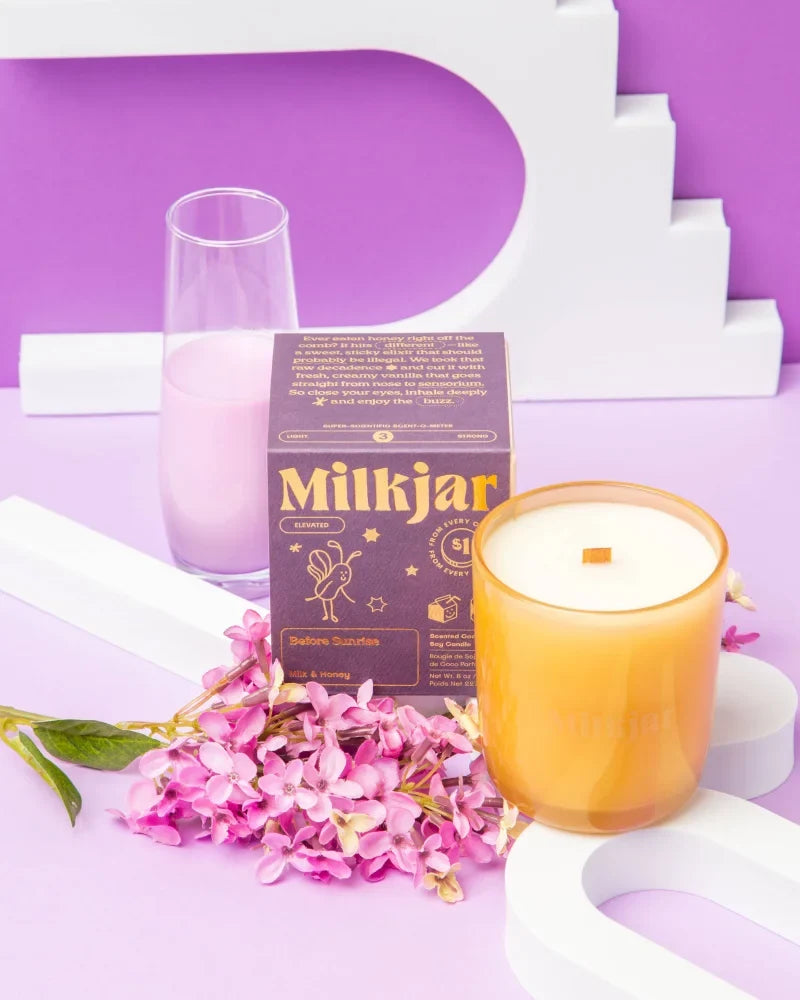 A golden amber coconut soy candle with wooden wick sits beside its before sunrise box and pink flowers by angus oliver