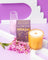 A golden amber coconut soy candle with wooden wick sits beside its before sunrise box and pink flowers by angus oliver