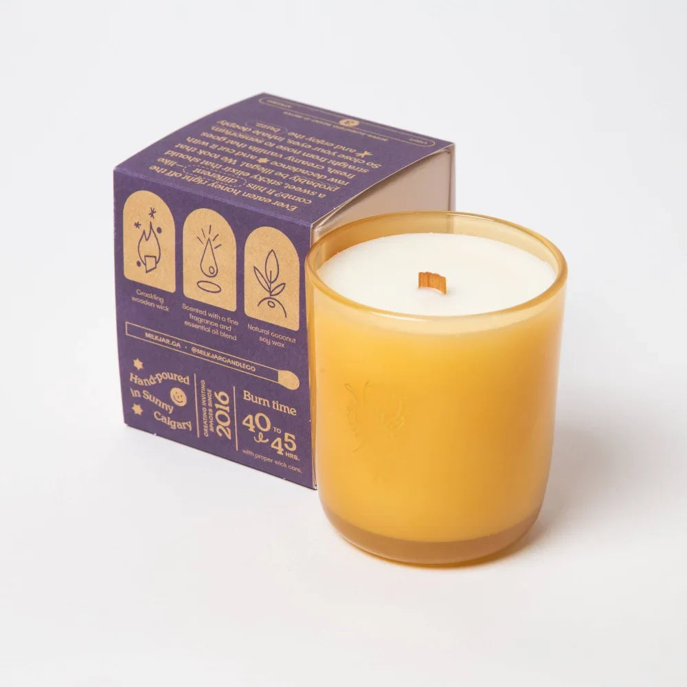 A golden amber glass candle with coconut soy wax and wooden wick next to its purple angus oliver box for cozy vibes