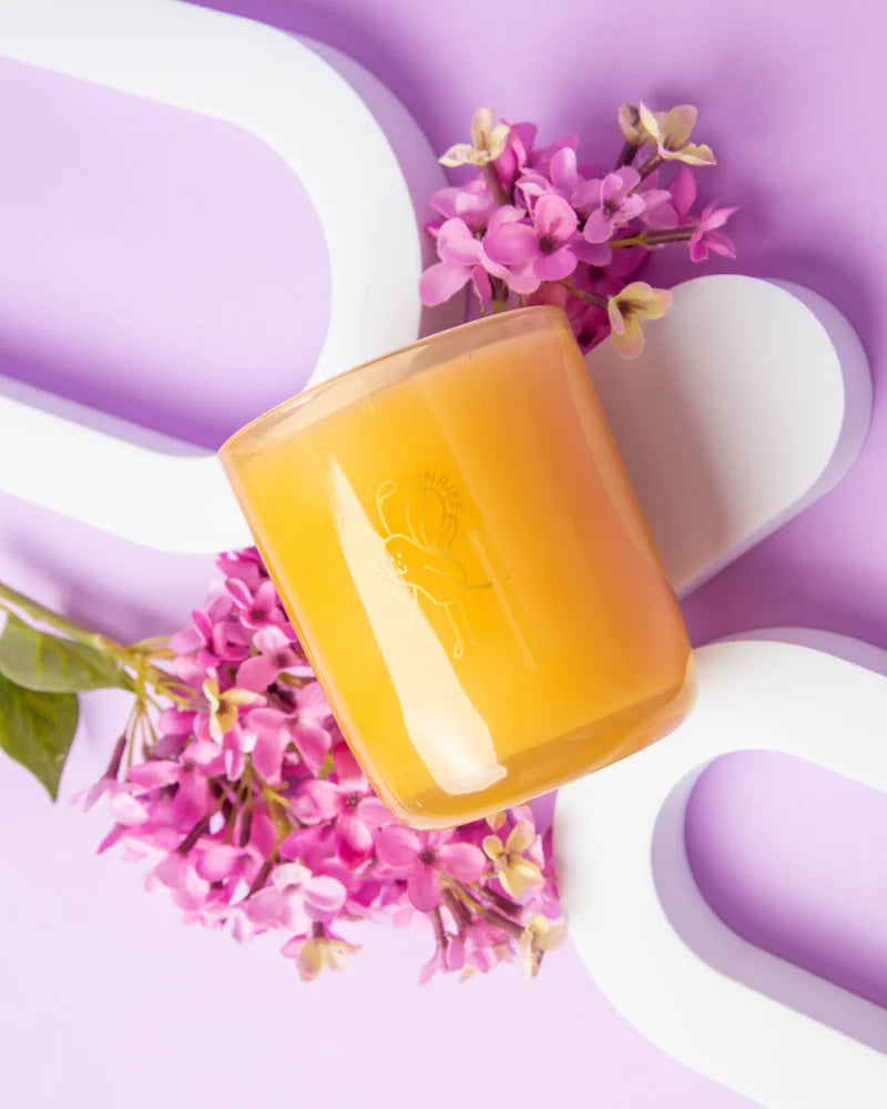 A translucent amber glass coconut soy candle jar with embossed logo surrounded by pink lilac blossoms from angus oliver