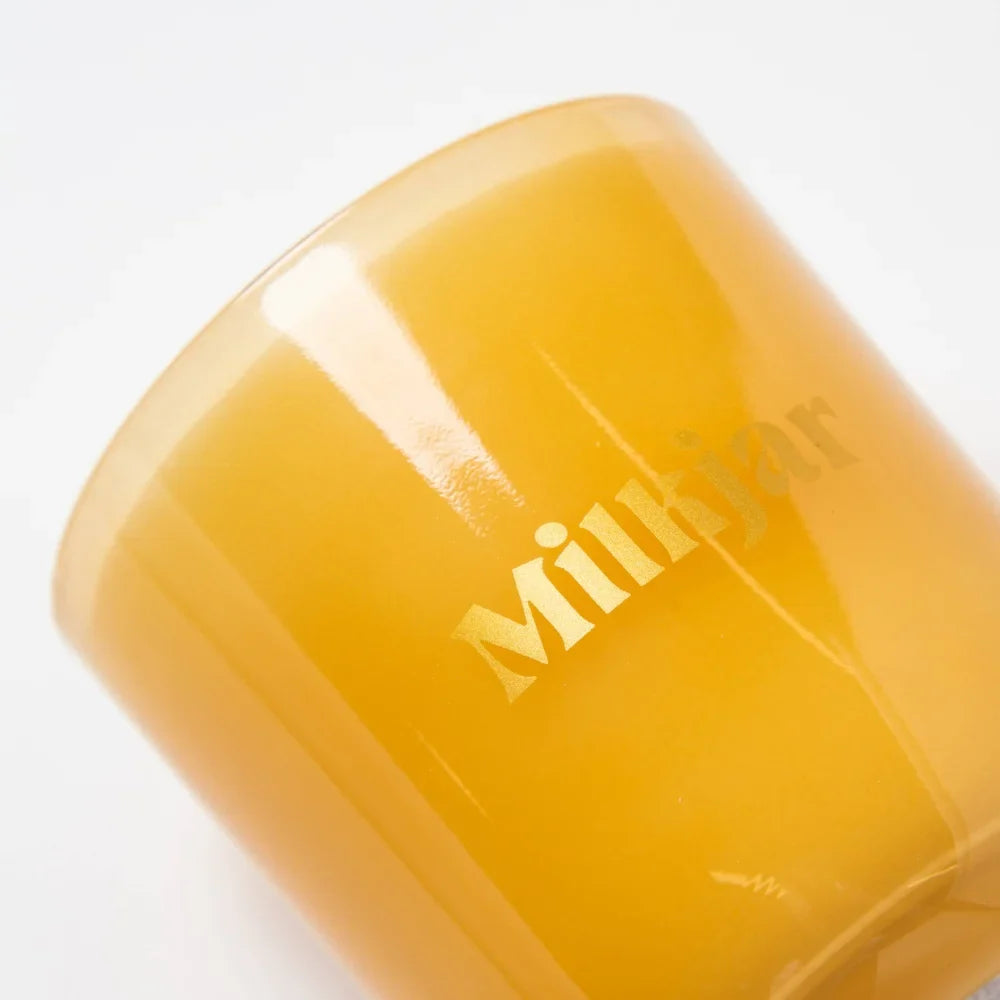 A translucent amber glass coconut soy candle jar with milkjar logo in gold lettering for cozy vibes like angus oliver rugs