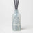 Translucent light blue glass reed diffuser bottle with etched teacup design holding gray sticks for blueberry tea scent
