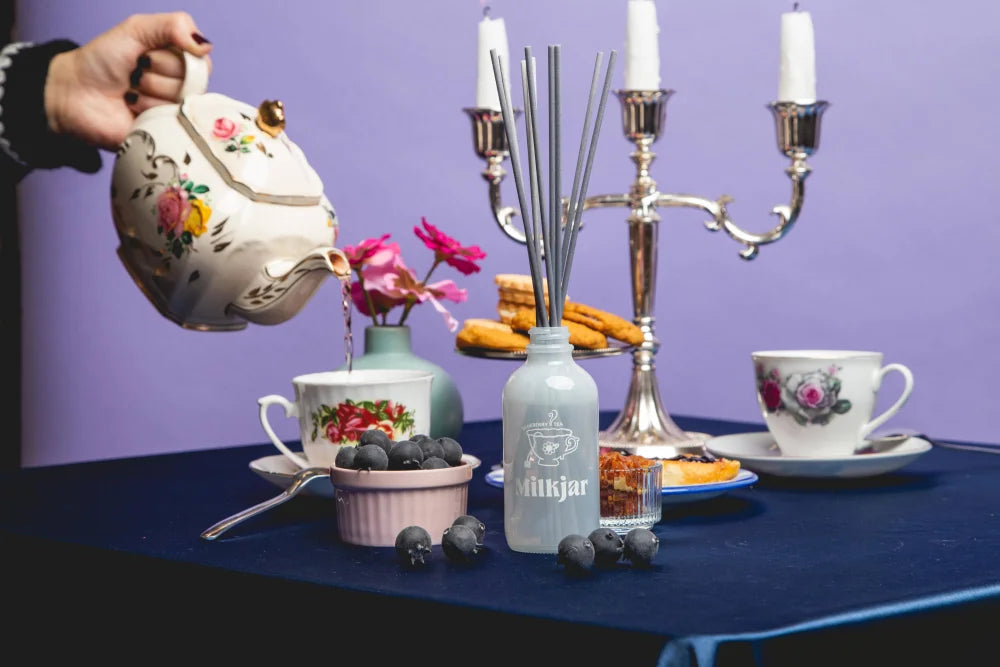 White ceramic teapot with floral design pouring blueberry tea over cup next to angus oliver blueberry tea reed diffuser