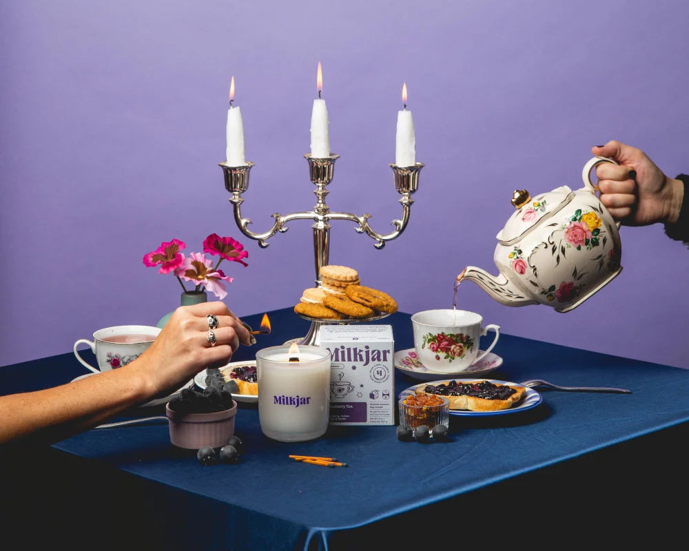 A hand pours from a white ceramic teapot with floral patterns beside a blueberry tea coconut soy wax candle