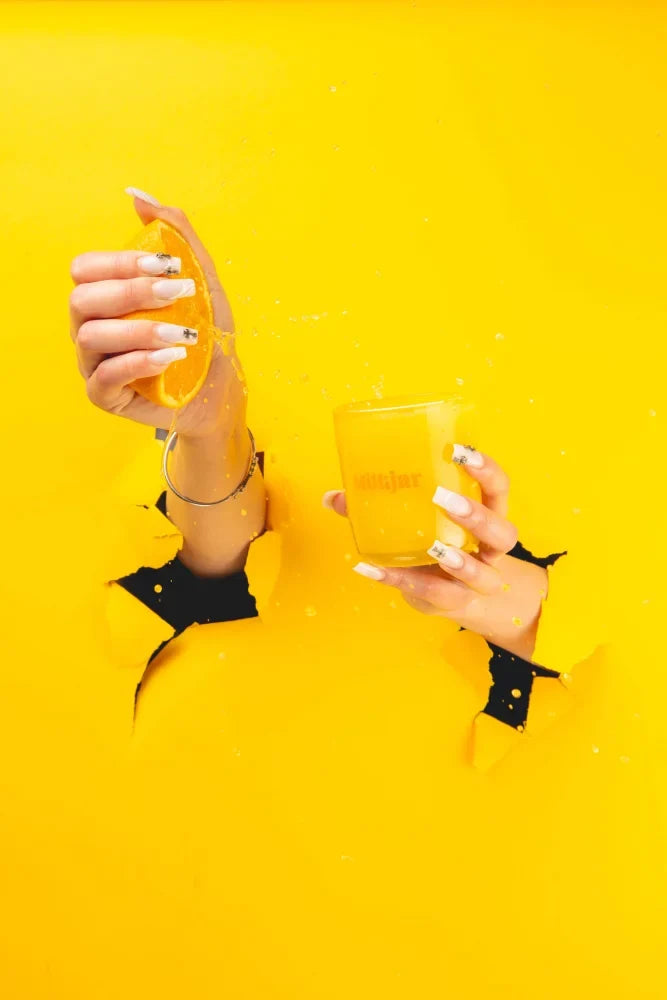 Hands with white and silver nail art squeeze an orange half juice splashes into a yellow glass near citrus essential oil coconut soy candle