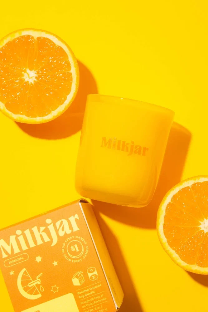 A vibrant orange citrus essential oil coconut soy candle in a clear glass jar with halved oranges on yellow background