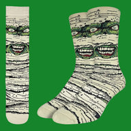 A pair of crew socks and a matching single sock shown flat against a white background. The socks feature a graphic print of a green monster face peeking through beige and black textured bandages, resembling a mummy.
