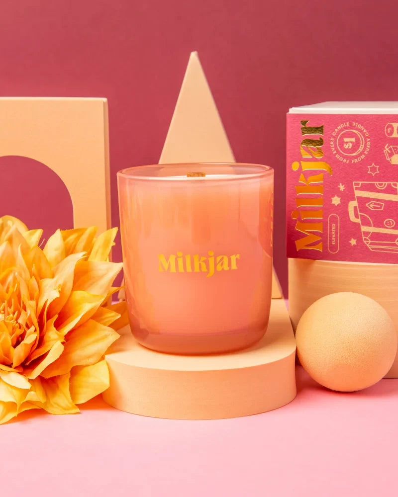 A peach-colored glass candle jar with a wooden wick and gold milkjar logo darjeeling patchouli and santal coconut soy blend