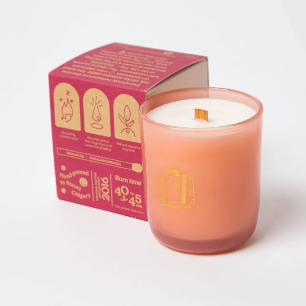 A peach-colored glass darjeeling patchouli and santal coconut soy candle with wooden wick next to its pink box