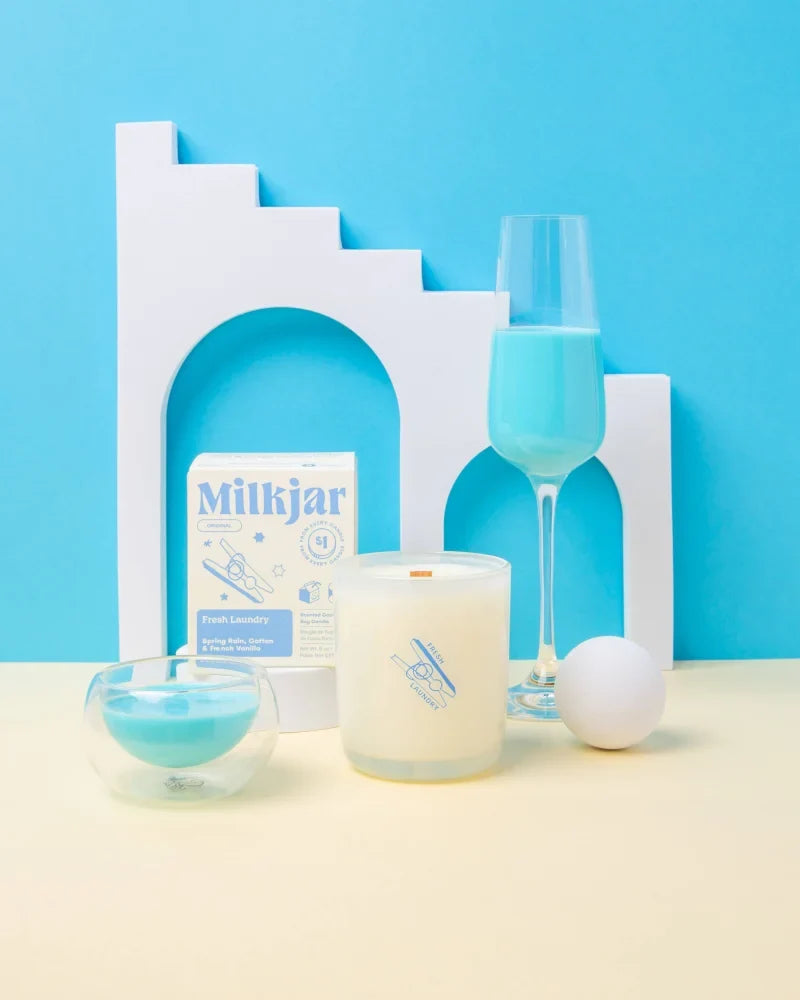 A creamy white fresh laundry rain cotton and vanilla coco soy 8oz candle in a glass jar with a minimalist blue logo