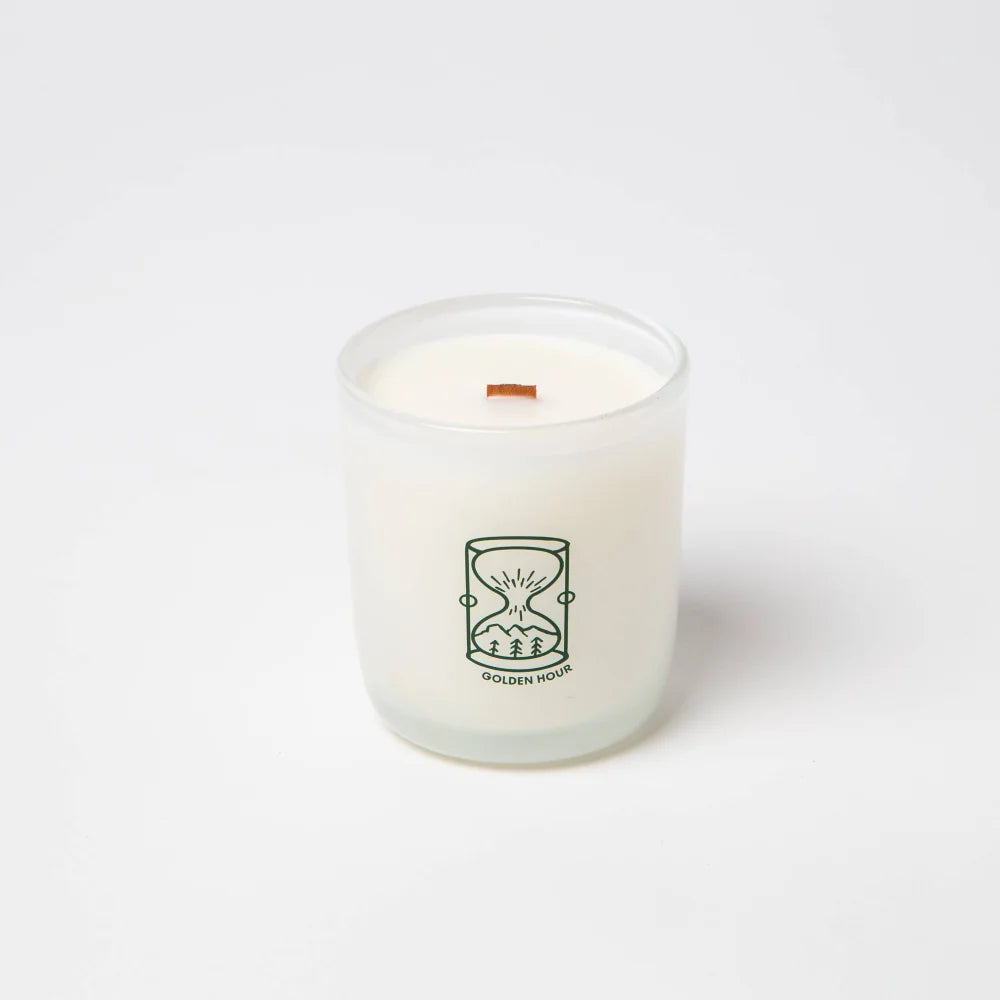 A white frosted glass candle jar with a green hourglass logo and wooden wick made from coconut soy wax