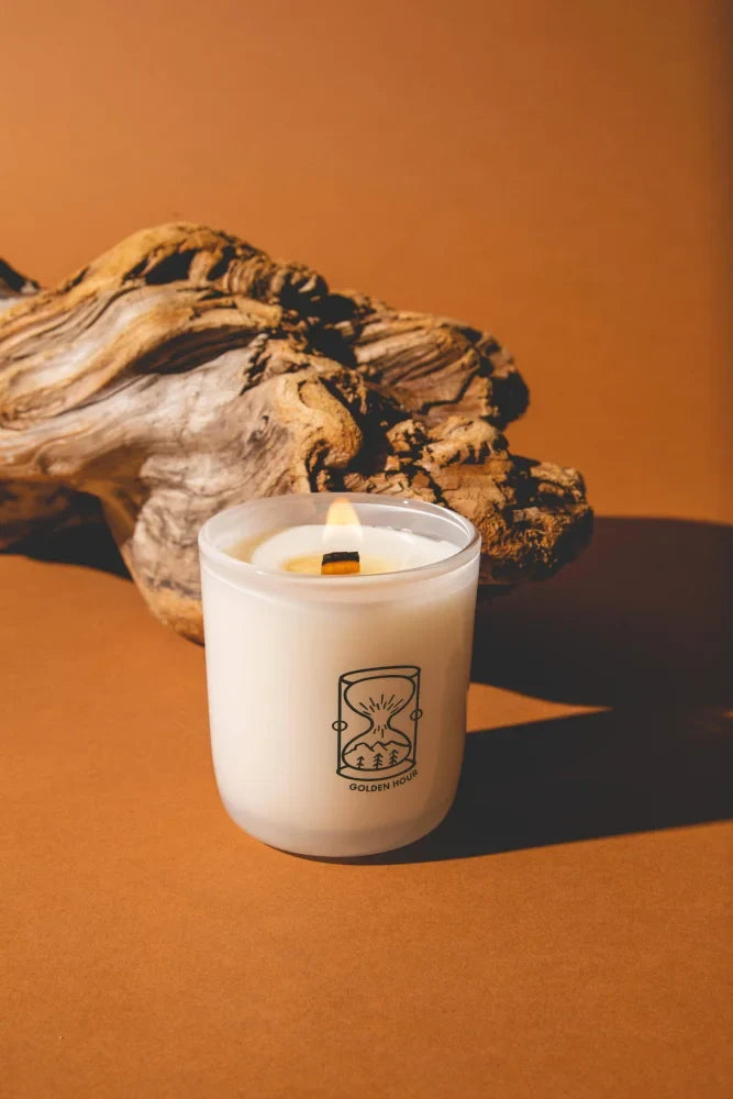A lit white candle in a clear jar with minimalist black hourglass logo golden hour made of coconut soy wax