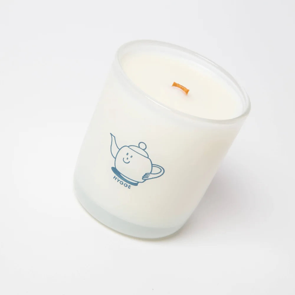 A white frosted glass hygge candle jar with a smiling teapot and orange wooden wick made from coconut soy