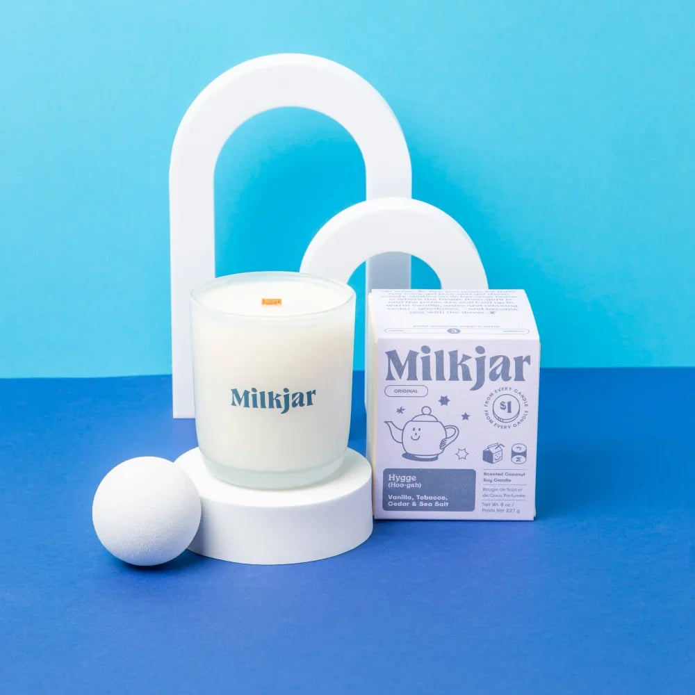 White milkjar candle in a clear glass jar with orange wick and teal logo hygge vanilla tobacco cedar coconut soy scent