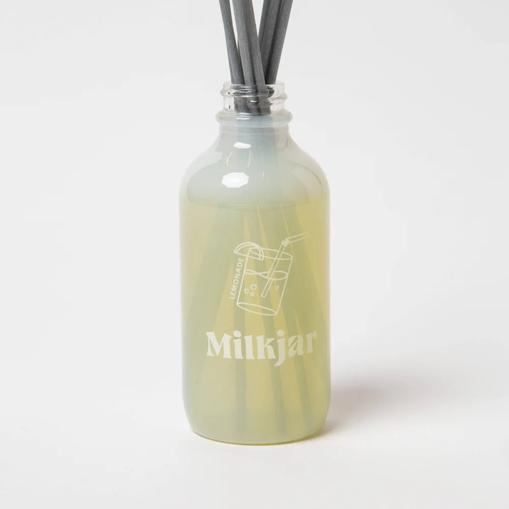 A translucent pale green glass reed diffuser bottle with minimalist white logo and dark gray sticks lemonadelime verbena pine