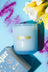 A light blue coconut soy candle in a frosted glass jar with gold lettering labeled moonrise neroli and plum