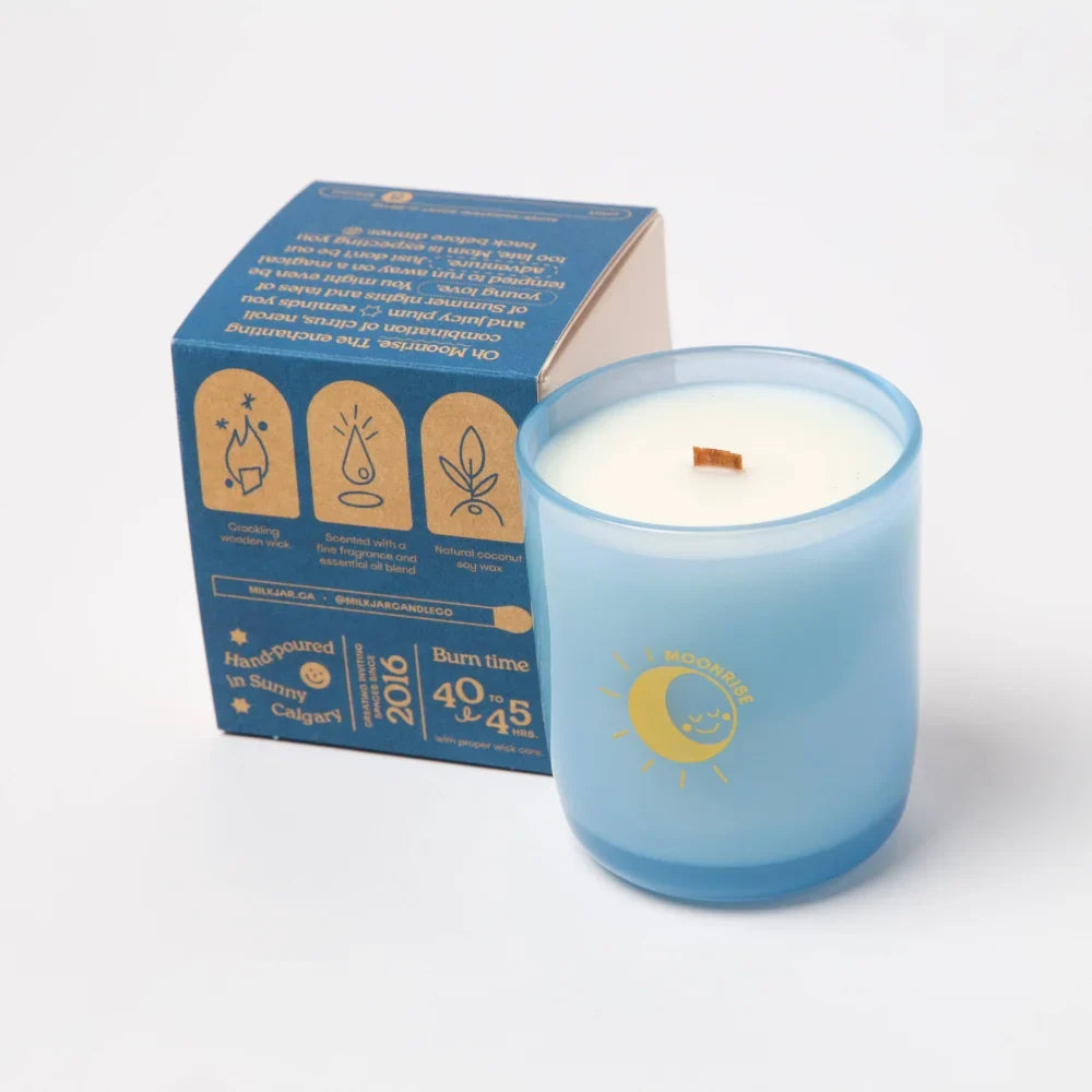 A light blue glass candle with wooden wick and sun-and-moon logo next to its box moonrise neroli plum coconut soy candle