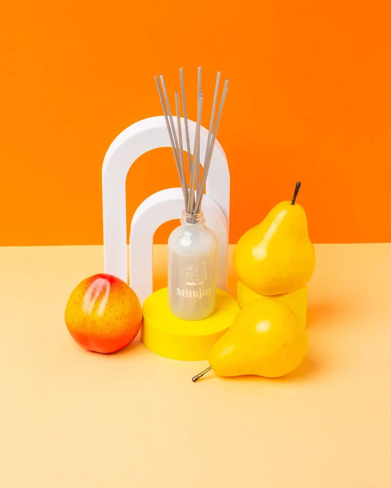 Clear glass sunnyside pear and nectarine 4oz reed diffuser bottle with silver reeds and minimalist logo on orange background