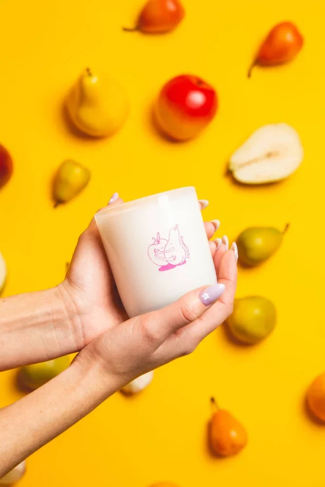 A white sunnyside coconut soy candle with pink apple and cat illustration held against a yellow fruit background smells fruity