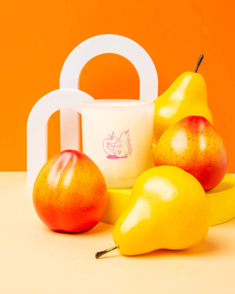 A creamy white coconut soy candle with a pink pear illustration nestled among yellow and red fruit sculptures
