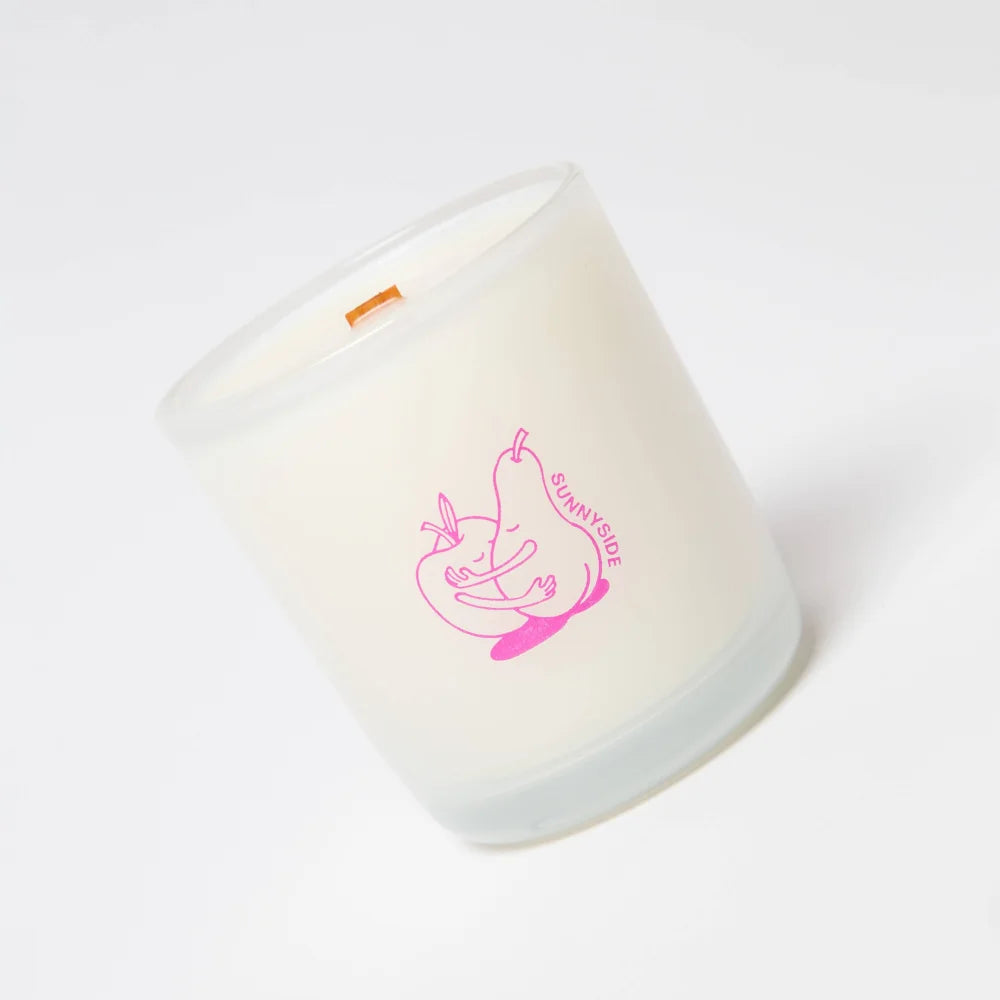 A frosted white glass coconut soy candle with a pink hugging pears design and sunnyside branding that candle smells fresh