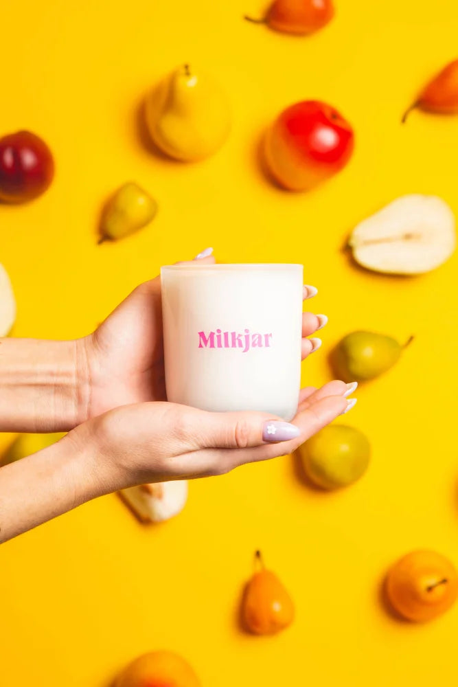 A white milkjar candle with pink lettering held in hands on yellow background featuring fruit, coconut soy candle smells