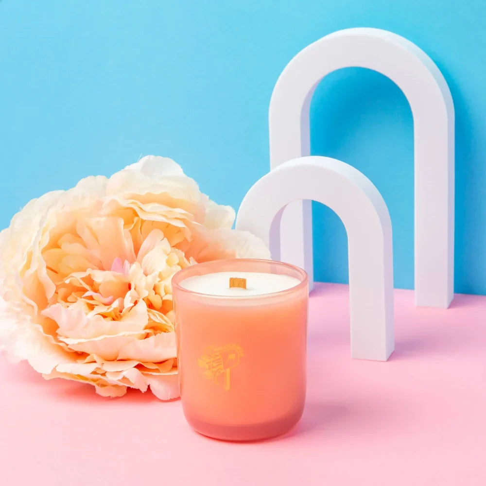 A peach-colored wallflower tobacco and peony coconut soy candle with wooden wick and gold foil branding