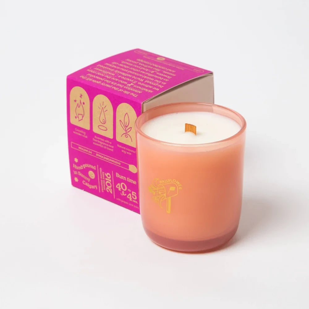A peach-colored coconut soy candle with a wooden wick and gold foil branding beside vibrant pink packaging