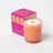 A peach-colored coconut soy candle with a wooden wick and gold foil branding beside vibrant pink packaging