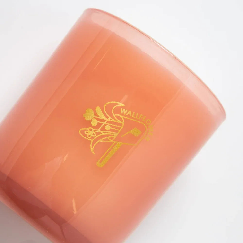 A peach-colored glass jar coconut soy candle with gold foil logo featuring floral design for wallflower tobacco peony
