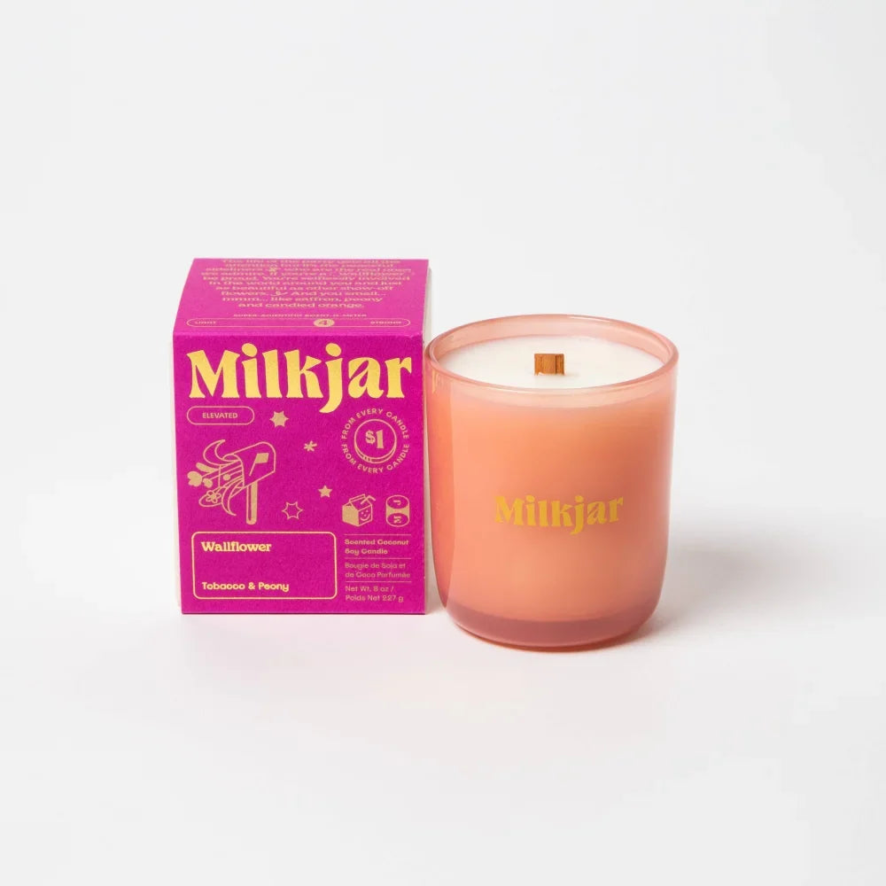 A peach-colored wallflower tobacco and peony coconut soy candle in frosted glass with gold lettering and wooden wick