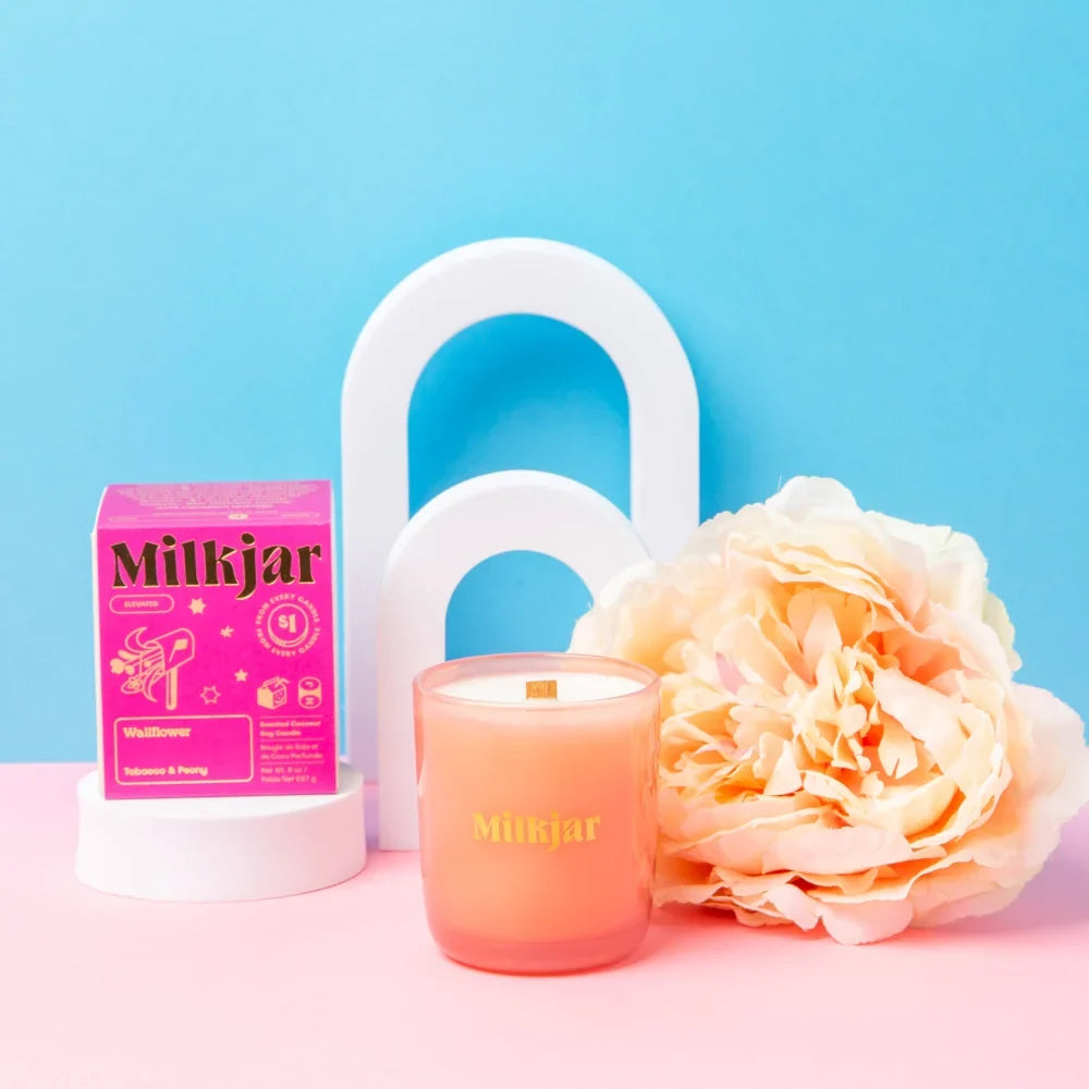 A peach-colored milkjar coconut soy candle with wooden wick sits next to its matching pink box wallflower tobacco and peony
