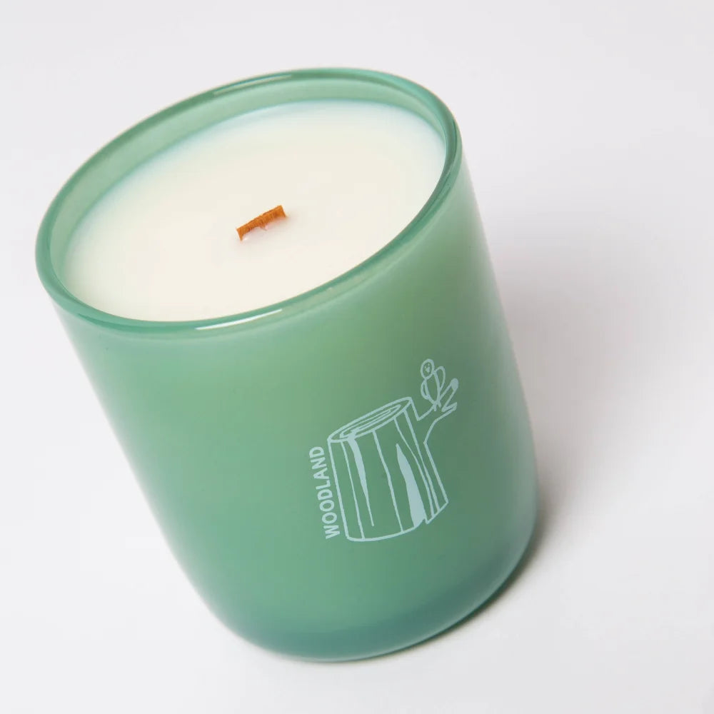 A matte green glass essential oil coconut soy candle jar with white wax and an orange wooden wick labeled woodland