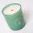 A matte green glass essential oil coconut soy candle jar with white wax and an orange wooden wick labeled woodland