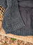 A cozy dark gray calgary cardigan made from zealand wool with a ribbed hem and textured weave on a basket