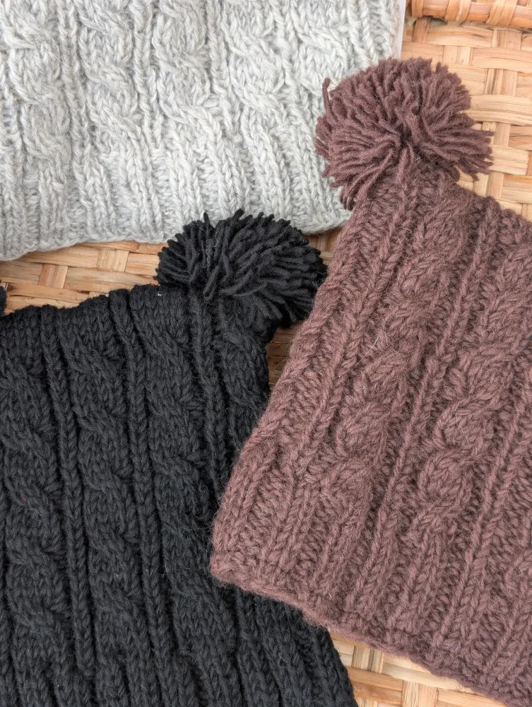 Three woolen heelah double pompom hats in gray black and brown with cable knit and fluffy double pompoms on top